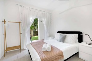 A bedroom with a bed, white curtains, a wooden rack, and a view of greenery outside.