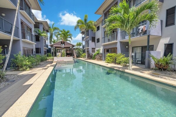 Una piscina con un pabellón, rodeada de edificios de varios pisos con balcones y palmeras.