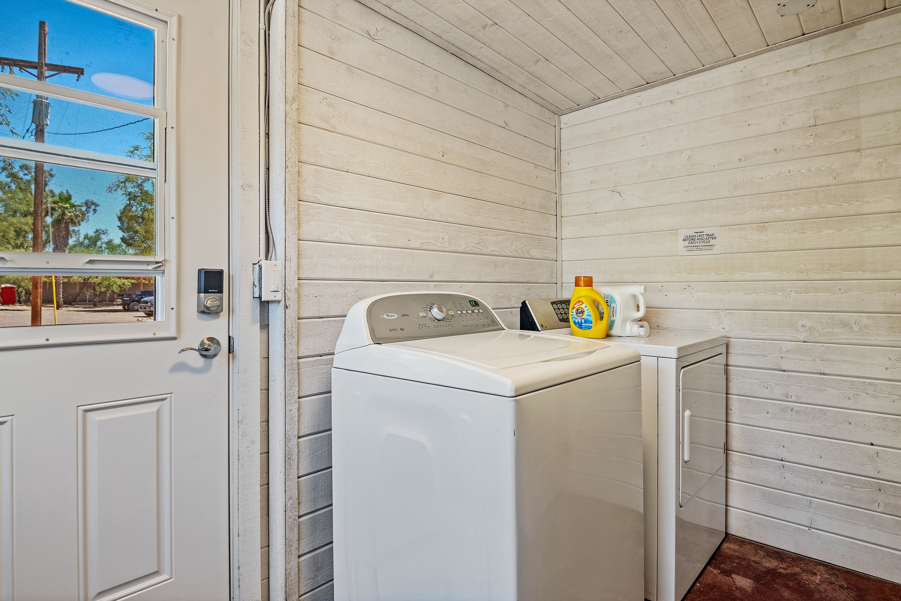 Une buanderie équipée d’un lave-linge et d’un sèche-linge à chargement frontal, d’une fenêtre donnant sur des arbres et une ligne électrique, et d’une porte avec une poignée.