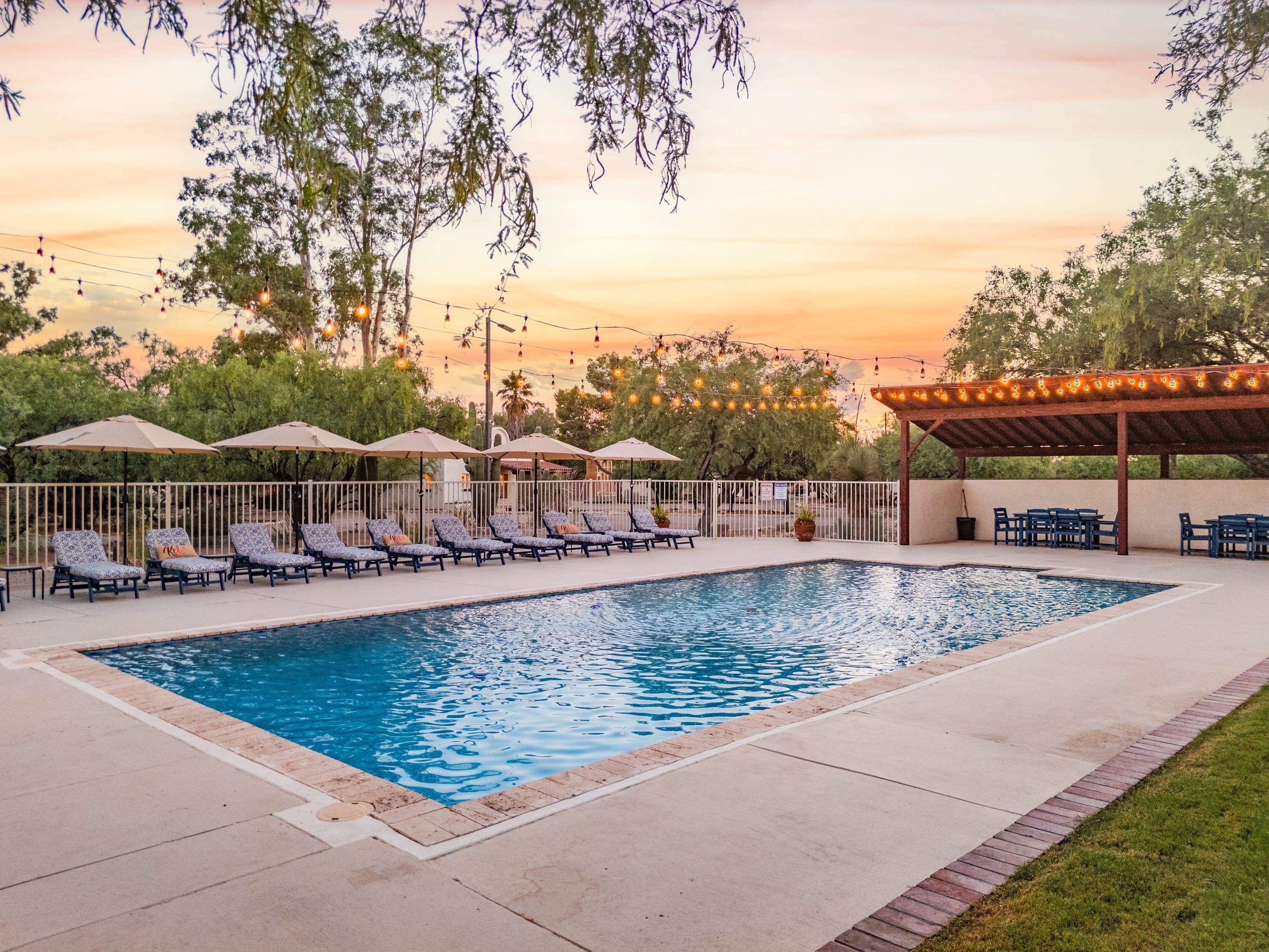 A rectangular pool with lounge chairs and umbrellas, surrounded by a fence and trees.