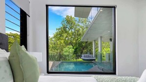 Ein modernes Schlafzimmer mit großem Fenster, Blick auf einen Swimmingpool und üppiges Grün.