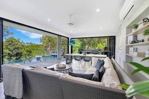Un espacio exterior moderno con una sala, una mesa de comedor y sillas, con vistas a una piscina y vegetación.