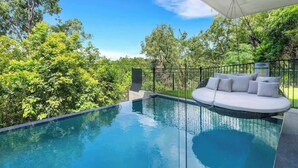 Ein unendlicher Pool mit einer weißen Sitzgelegenheit im Freien und üppiger Bepflanzung im Hintergrund.
