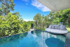 Una piscina infinita con un área de estar exterior blanca y abundante vegetación al fondo.
