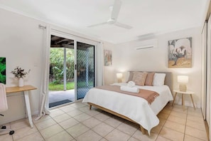 Ein Schlafzimmer mit Bett, Nachttischlampen, Deckenventilator, einer zur Balkontür führenden Tür und einem kleinen Tisch mit einer Vase.