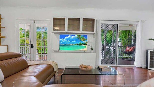 A living room with a leather sofa, a glass coffee table, a TV on the wall, and a sliding glass door leading to an outdoor area. - Tropical Refuge—Classic Queenslander Paradise (Aeroglen)