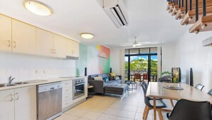 A modern kitchen with a dishwasher, oven, and a dining area with a table and chairs. - The Loft—Trendy Coastal Charm with Pool (Cairns North)