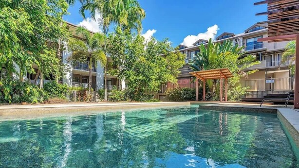 A clear swimming pool surrounded by lush greenery and multi-story buildings. - The Loft—Trendy Coastal Charm with Pool (Cairns North)