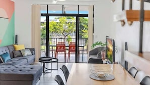 A modern living room with a sofa, dining table, chairs, and a balcony with red chairs. - The Loft—Trendy Coastal Charm with Pool (Cairns North)
