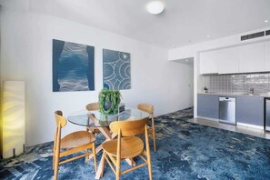 Une pièce moderne avec une table en verre, des chaises en bois, un coin cuisine et des œuvres d’art abstraites accrochées au mur.