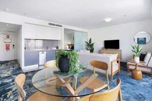 Une chambre d’hôtel moderne avec une table à manger en verre, des chaises en bois, un canapé, une télévision et une grande plante en pot.