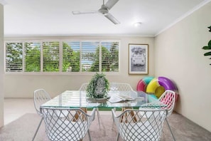 A dining area with a glass table, white chairs, a ceiling fan, a window with blinds, and a framed picture on the wall.