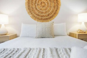 Un letto con copripiumino bianco, un cuscino decorato e un pannello decorativo appeso al muro. Ci sono due comodini, ognuno con una lampada e un piccolo oggetto decorativo.
