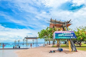 Una spiaggia con un cartello che riporta 'CAIRNS ESPLANADE', un'area piscina e turbine eoliche sullo sfondo.