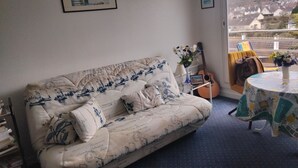 A cozy living room with a patterned sofa, a guitar, a vase with flowers, and a view of houses through a window.