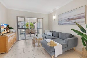 A living room with a grey sofa, a television, a dining area with chairs and a table, and a large window.