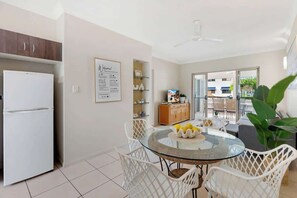 Une cuisine moderne avec des meubles blancs, un réfrigérateur blanc, une table à manger en verre avec des chaises blanches, et un panneau d’accueil accroché au mur.