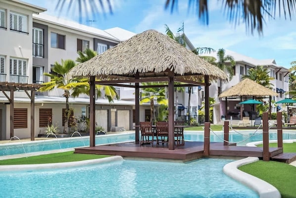 A pool area with a thatched gazebo, lounge chairs, and a poolside view of a resort building.