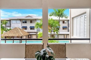 A balcony with a view of a pool, a thatched-roof structure, and a building with multiple windows.