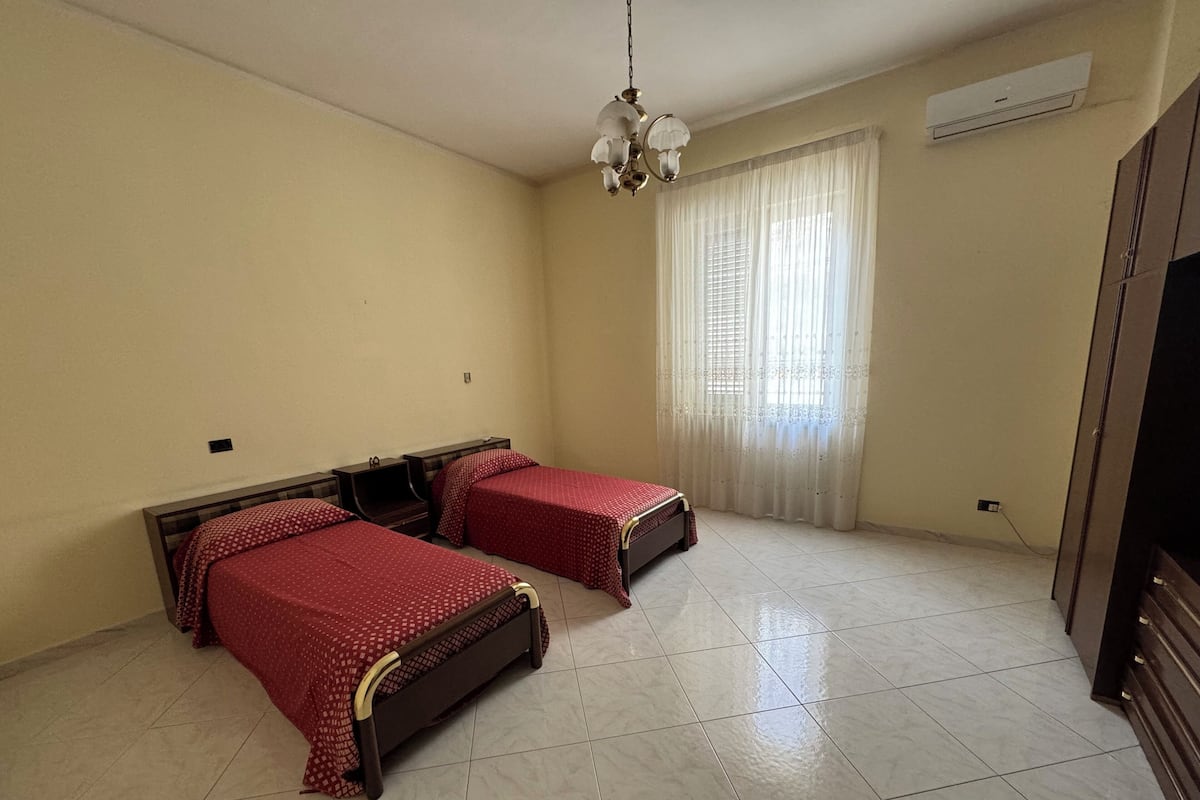 A room with two beds, a ceiling light, a window with curtains, and an air conditioning unit.