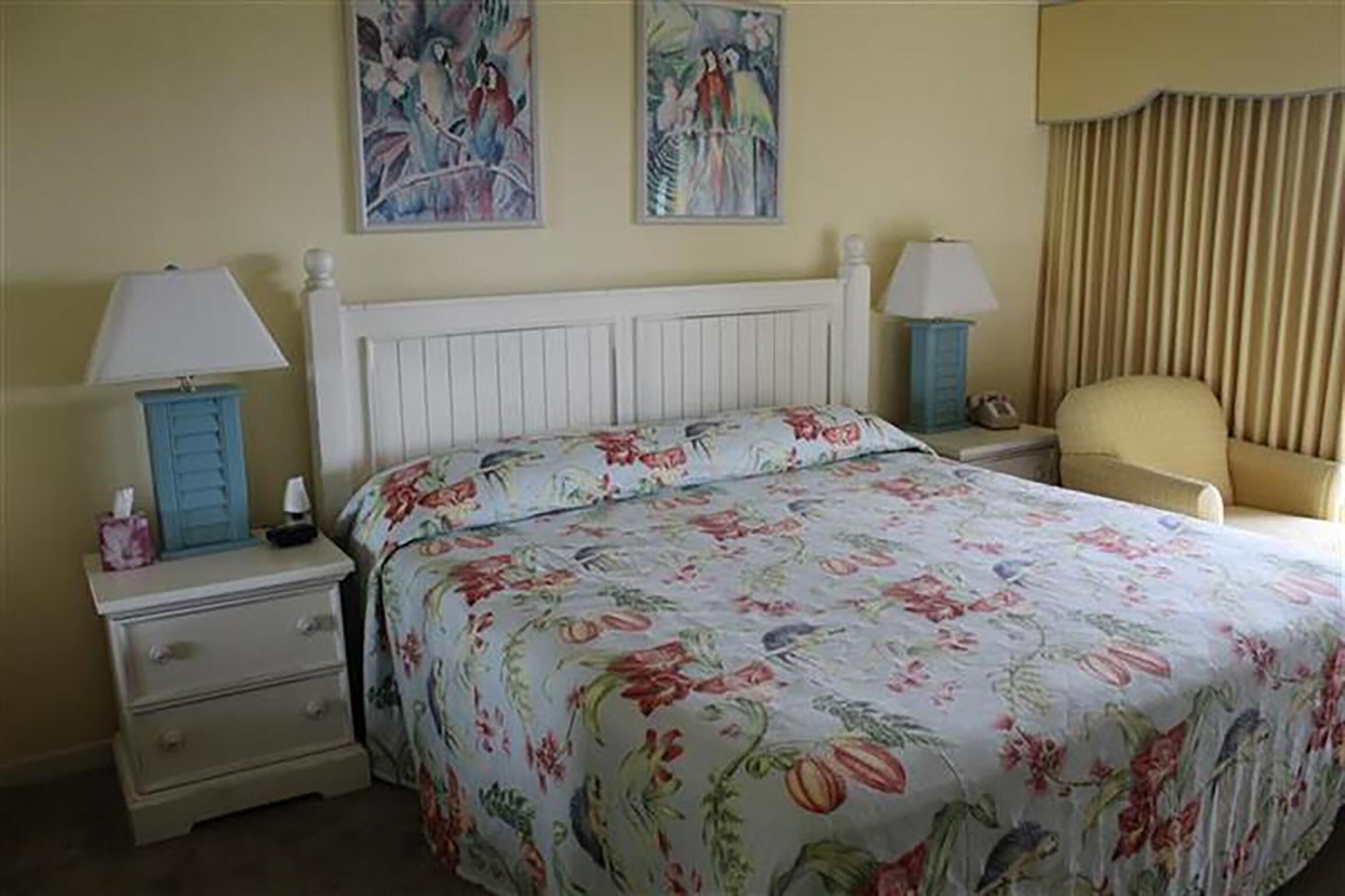 Une chambre à coucher avec une couvre-lit à motifs floraux, une tête de lit blanche, deux tables de chevet avec des lampes, une chaise et deux tableaux accrochés au mur.