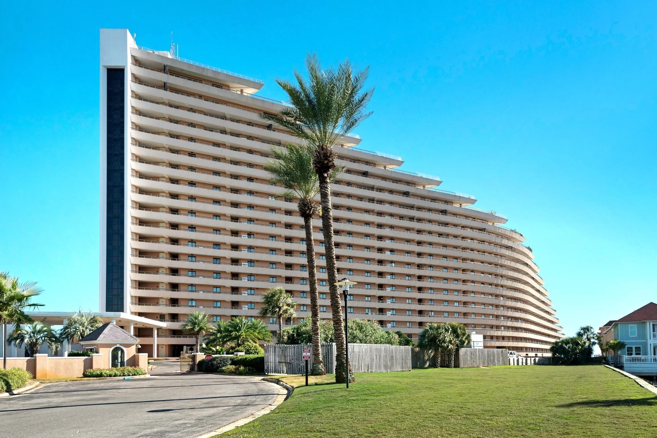 A large, multi-story hotel with a curved facade, a central entrance, and a well-maintained lawn with palm trees.