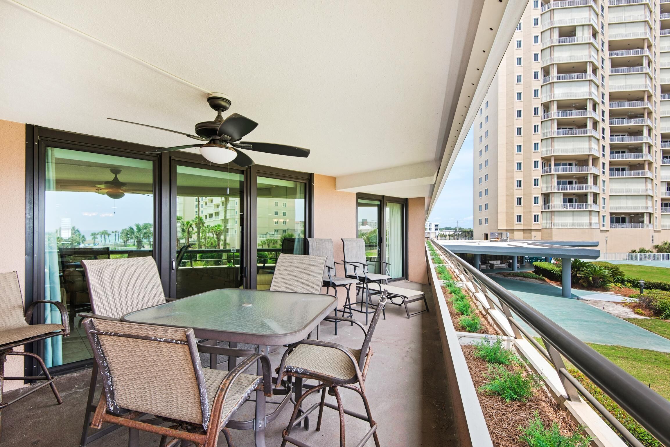 A balcony with a glass table and chairs, a ceiling fan, and a view of a high-rise building and greenery.
