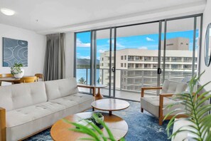 A modern hotel room with a sofa, two armchairs, a coffee table, and a large window offering a view of a building and mountains.