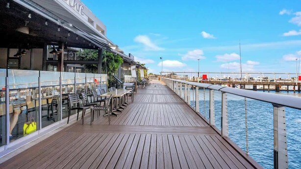 A wooden deck with outdoor seating, a glass railing, and a view of the water. - City Living at Harbour Lights on the Waterfront (Cairns)