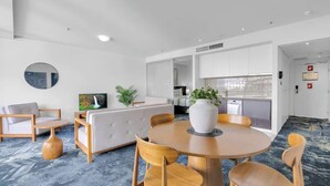 A modern hotel room with a round dining table, wooden chairs, a sofa, a TV, and a kitchen area. - City Living at Harbour Lights on the Waterfront (Cairns)