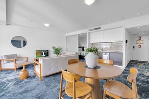 A modern hotel room with a round dining table, wooden chairs, a sofa, a TV, and a kitchen area.