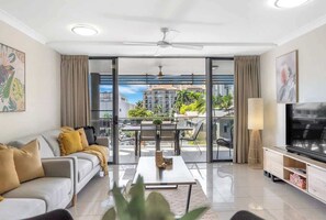 Ein modernes Wohnzimmer mit Sofa, Couchtisch, Fernseher und Balkon mit Blick auf Gebäude und Grünflächen.