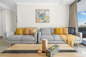 Un salon avec deux canapés gris, des coussins jaunes, une table basse en bois, un jeu de puzzle et un tableau coloré accroché au mur.