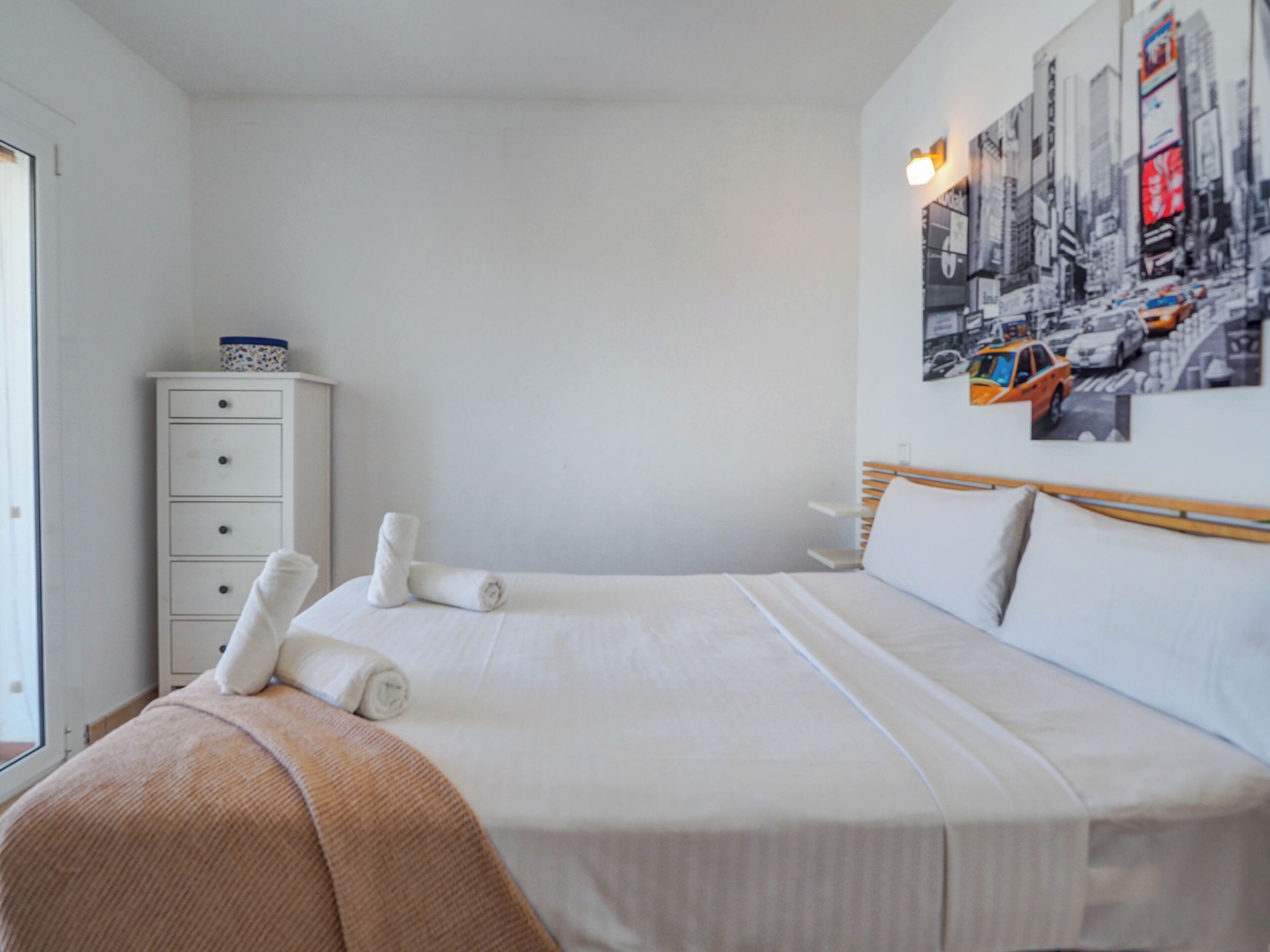 Un lit bien fait, avec du linge de lit blanc et un plaid beige, une commode blanche avec des tiroirs, et un tableau représentant un paysage urbain accroché au mur.