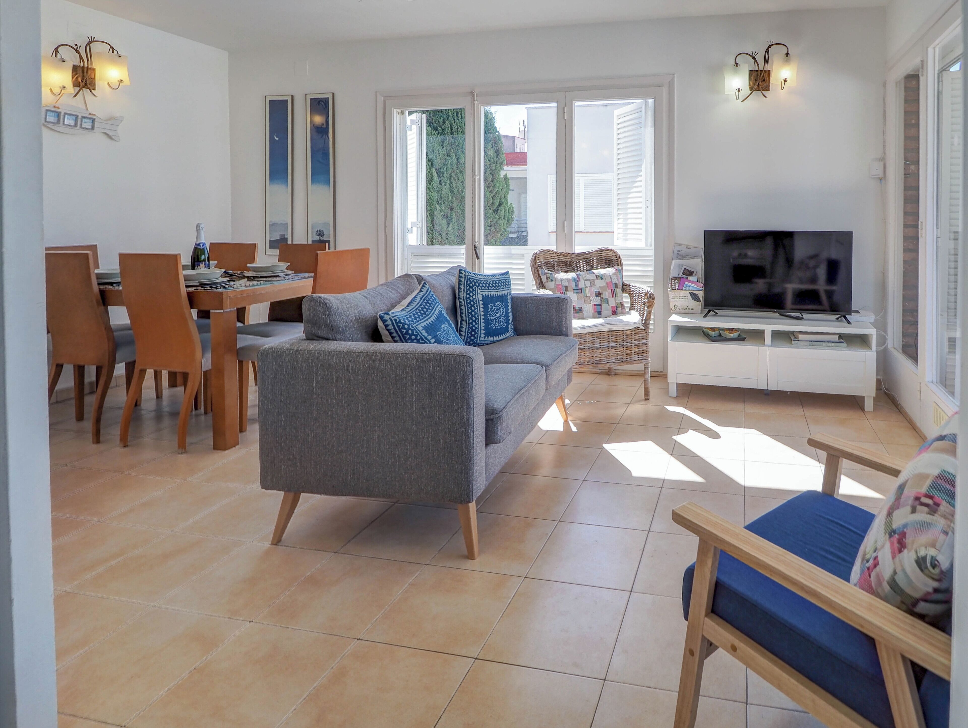 A living room with a grey sofa, a dining table with chairs, a TV on a white stand, and large windows with blinds.