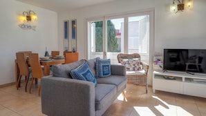 Un salon avec un canapé gris, une table à manger avec des chaises, un téléviseur posé sur un meuble et une fenêtre avec des stores.