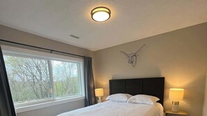 A bedroom with a large bed, two bedside lamps, a ceiling light, a window with curtains, and a wall-mounted decor piece.