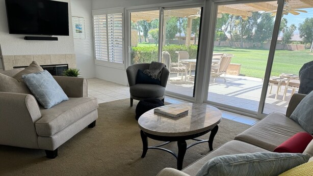 A living room with a sofa, armchair, and coffee table, large windows with a view of a backyard. - Beautiful and charming condo in Woodhaven Country Club! Recently updated (Palm Desert)