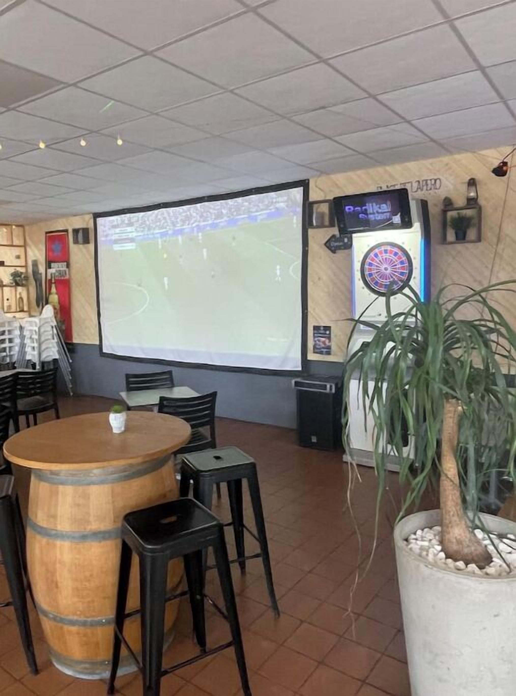 Una habitación con una pantalla grande mostrando un evento deportivo, un dardo y una mesa de barrica de madera con sillas.