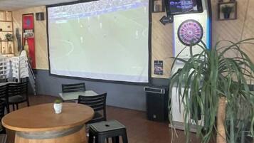 Una habitación con una pantalla grande mostrando un evento deportivo, un dardo y una mesa de barrica de madera con sillas.