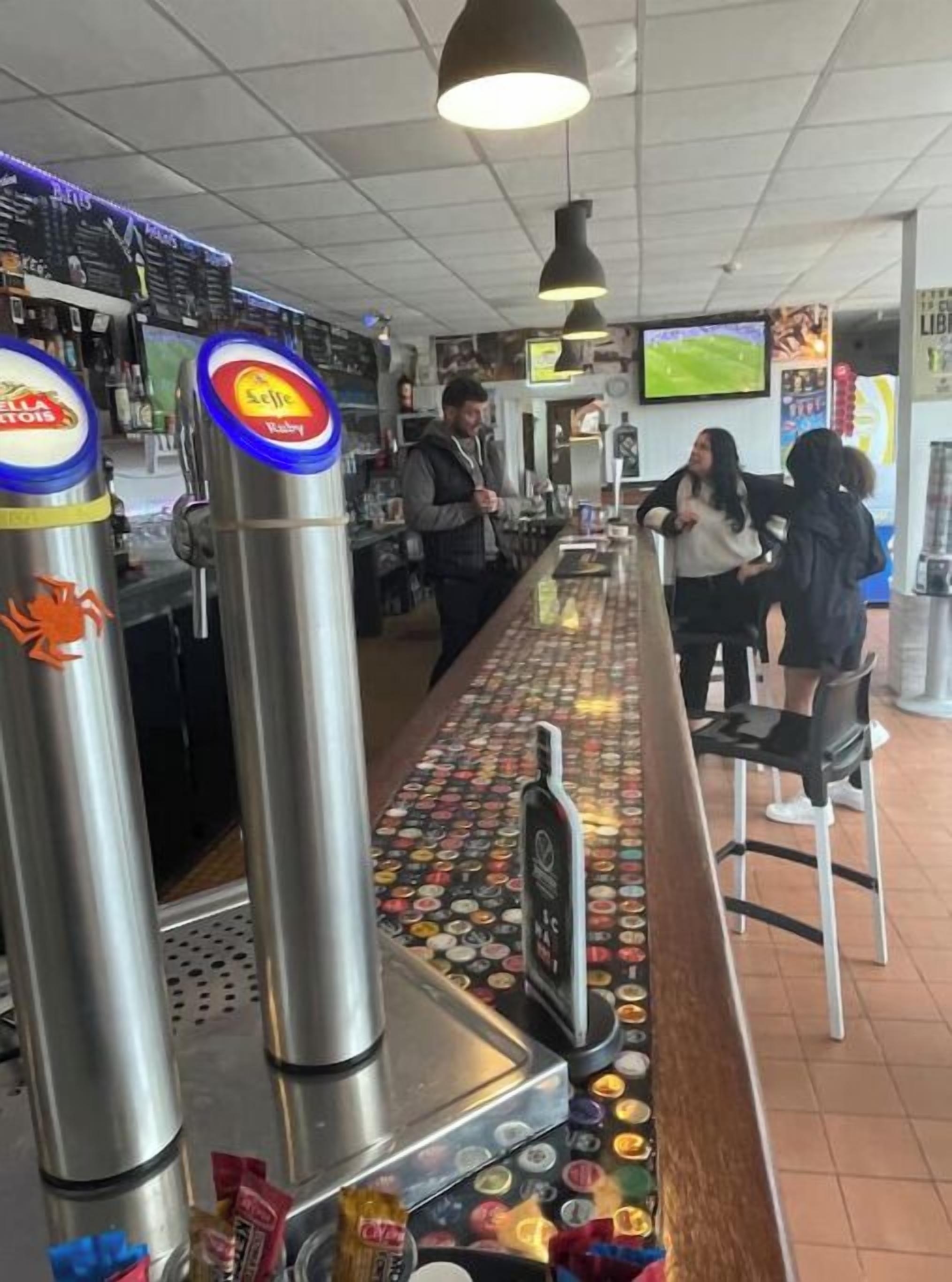 Un espace bar doté de comptoirs en acier inoxydable, d’une variété de bouteilles et d’un sol carrelé orné de capsules de bouteilles.