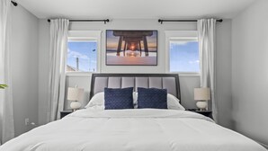 Een net opgemaakt bed met witte beddengoed en twee blauwe kussens, met nachtkastjes met lampen ernaast, in een kamer met een groot raam en een zonsondergangschilderij boven het bed.