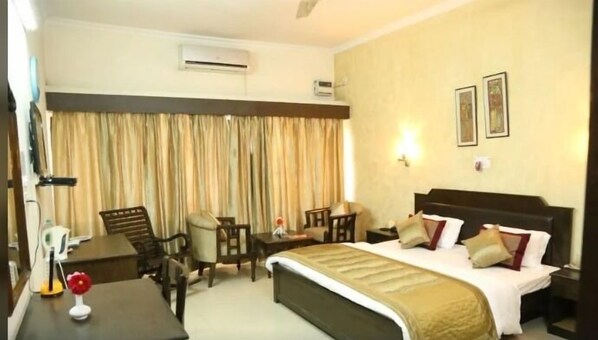 A hotel room with a bed, a desk, a chair, and a table. There are curtains on the window, and an air conditioning unit is mounted on the wall. - Badkhal Lake Resort (Faridabad)