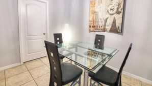 Een eetruimte met een glazen tafel en zwarte stoelen, een witte deur en een poster van Marilyn Monroe aan de muur.