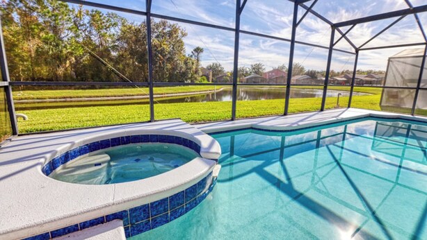 Een zwembad met een jacuzzi, omgeven door een overdekt terras met uitzicht op een grasveld en een waterpartij.