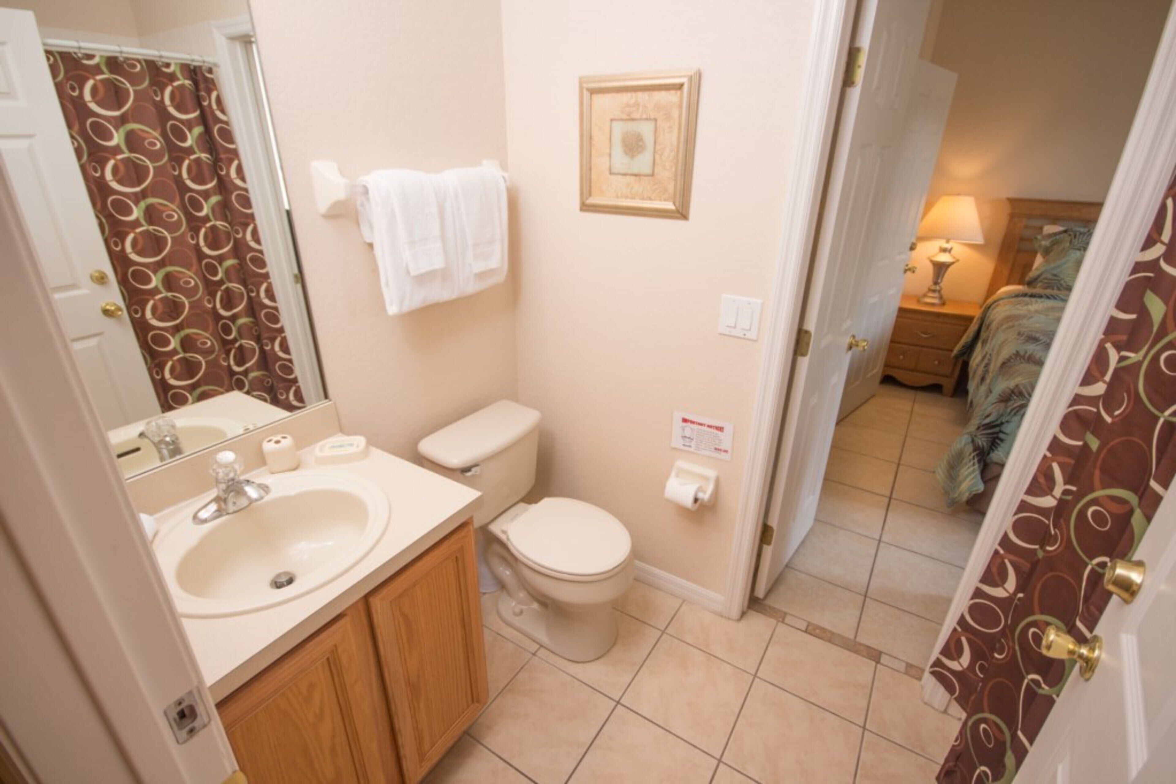 A bathroom with a sink, toilet, and mirror. There is a towel rack with towels, a framed picture on the wall, and a door leading to another room.