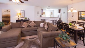 A living room with a sectional sofa, a dining table with chairs, a ceiling fan, and a kitchen in the background. - Robert's Cypress Pointe Vacation Home (Davenport)