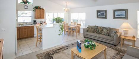 A spacious living area with a sofa, a coffee table, and a dining table with chairs. There is a kitchen area with wooden cabinets and a window.