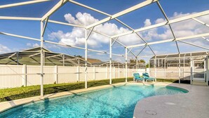 Ein überdachter Pool mit einer klaren, blauen Wasseroberfläche, umgeben von einem weißen Zaun und einer überdachten Sitzgruppe mit zwei Liegestühlen.