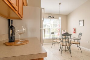 A kitchen with a wine bottle and glass on the counter, a dining area with chairs and a table, and a window with blinds.